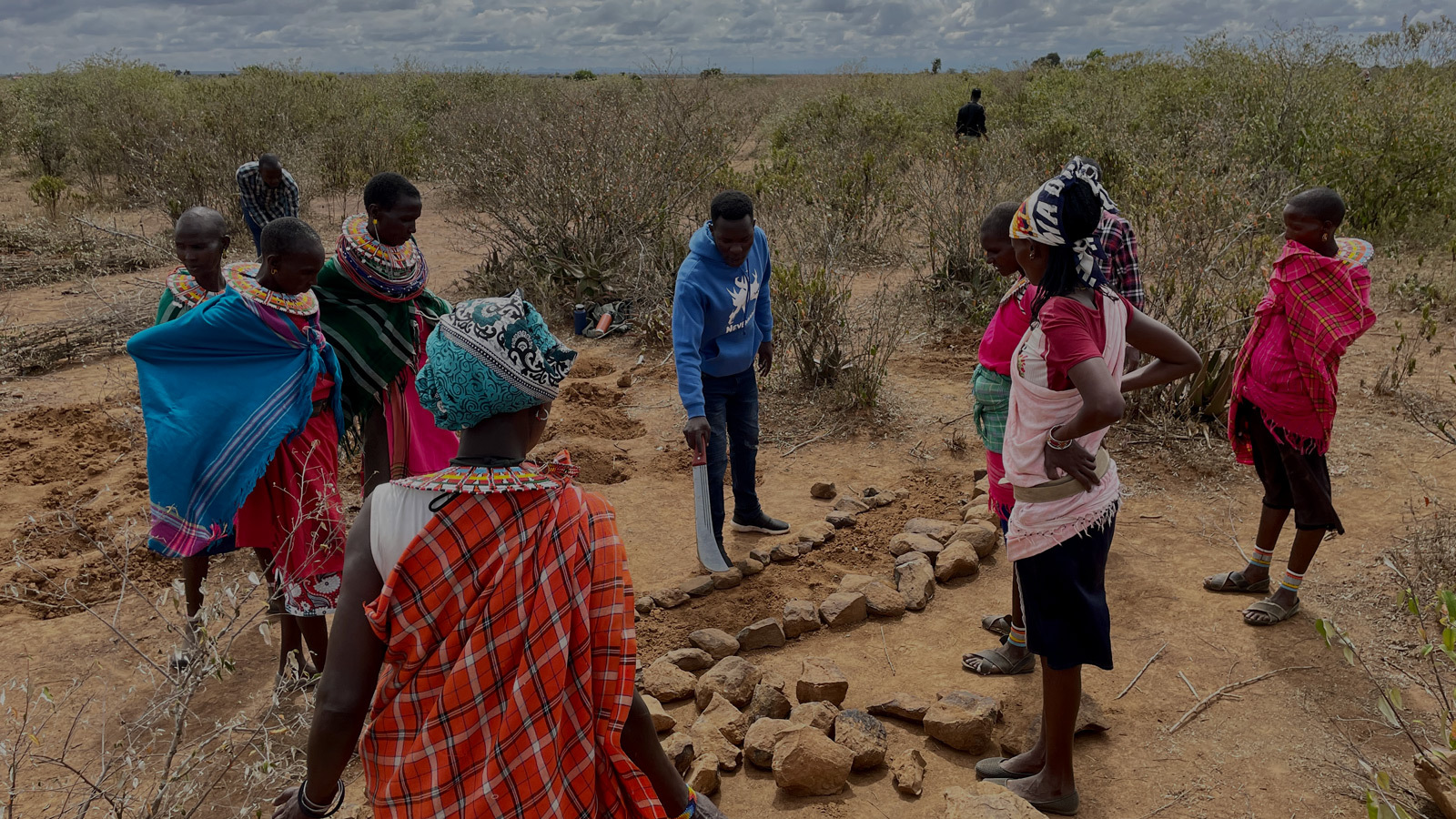Group standing in dry grasslands, pointing at water harvesting structure.